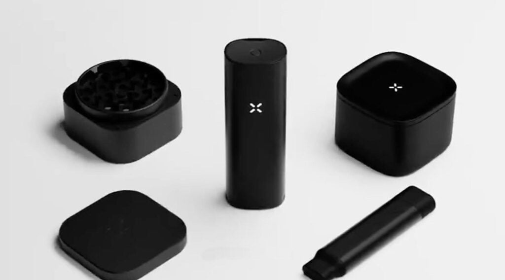 **Alt text:**
Black PAX vaporizer accessories arranged on a white background, including a portable vaporizer device, a grinder, a storage container, a lid, and a loading tool. The sleek matte-black accessories feature the illuminated PAX logo.