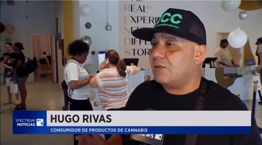**Alt text:**
News interview screenshot showing a man wearing a black cap with a green logo speaking indoors at a cannabis-related event. Several attendees are visible in the background, and a lower-third graphic identifies him as Hugo Rivas, a cannabis consumer, during a Spectrum Noticias NY1 segment.