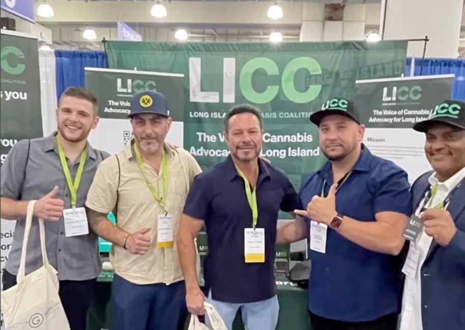 **Alt text:**
Group photo of five attendees standing in front of a Long Island Cannabis Coalition (LICC) booth at a cannabis industry expo. The participants wear event badges and LICC-branded hats, posing together in front of banners promoting cannabis advocacy on Long Island.